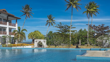 Blue resort swimming pool. Sun beds under a canopy, curtains fluttering in the wind. Green tropical vegetation. Tall palm trees against the sky and clouds. A hotel building. The ocean is far away. 