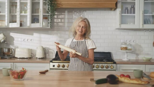 Carefree old woman enjoying music and dancing alone in home kitchen, portrait. Charming elderly lady singing into wooden rolling-pin ,good mood and positivity, anti-stress and recharge with energy