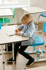 A schoolboy sits at a desk and writes with a pen in a notebook, at school, in the classroom
