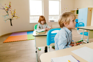 Obraz premium A schoolboy sits at a desk and looks in front of him while holding pencils, a classmate sits behind him at a desk, at school, in the classroom