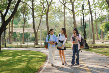 Fototapeta premium Three female university students carrying backpacks and books are walking and talking together at a park