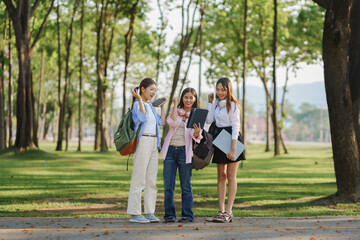 Obraz premium Three young women celebrating good news on a tablet, holding backpacks and books while enjoying a sunny day in the park