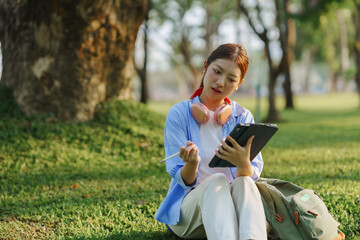 Young woman sitting on grass using digital tablet and stylus with puzzled expression, backpack and...