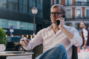 Young freelancer in white shirt making vibrant phone conversation, multitasking outdoors, sitting...