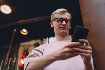 Young trendy man browsing smartphone outdoors, relaxed urban setting, wearing glasses and pastel casual clothing, modern communication, technology integration into daily street lifestyle