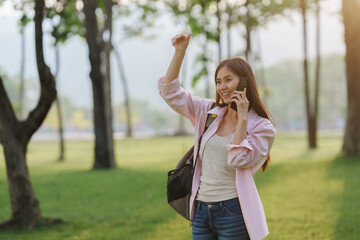 Fototapeta premium Happy student waving and chatting on a smartphone while enjoying a sunny day in the park, surrounded by nature and greenery