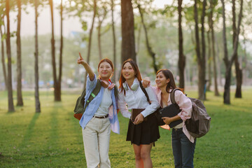 Three young female students with backpacks and books walking together in a park, one of them pointing to something in the distance