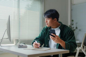 Asian freelancer working from home taking notes while looking at computer and holding smartphone