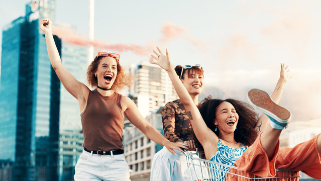 Friends, girls with trolley in city laughing for funny adventure, playing and bonding on sidewalk. Excited, color smoke or women pushing cart in urban town with freedom, confidence and weekend break