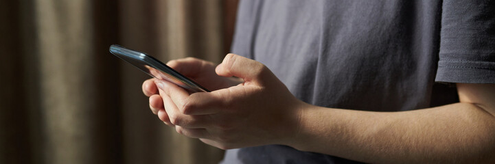 Header of hands of person holding smartphone while standing indoors next to curtain