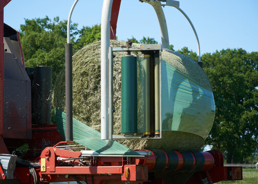 Round bale of hay being wrapped in green silage stretch film coming from a large roll rotating around it