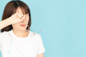 Asian​ woman​ in eyeglasses rubbing tired eyes relief vision pain after work over blue background.​ Eye strain symptoms include watery eyes, dry eyes, blurred vision and itching eyes.