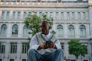 Obraz premium stylish Black male sits outside a historic building, holding his smartphone with a thoughtful expression.Young man modern outfit and confident posture reflect ambition and a digital lifestyle