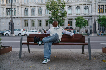 Obraz premium stylish Black man sits on bench in urban setting, casually using his smartphone. appears relaxed, possibly browsing social media or handling work remotely, with modern cityscape in background