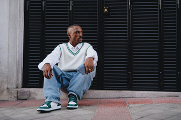 Young African American man in casual streetwear sitting outdoors, looking to the side with a thoughtful expression. Black shutters create a modern urban backdrop. Holding smartphone, relaxed pose