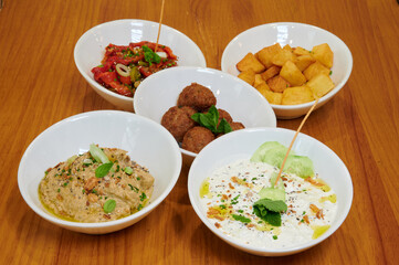 A wooden table is set with five white bowls filled with various dishes. The bowls contain hummus, tabbouleh, potato wedges, and other food items