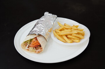 A wrap with lettuce and tomatoes sits on a white plate next to a plate of french fries. The wrap is wrapped in foil