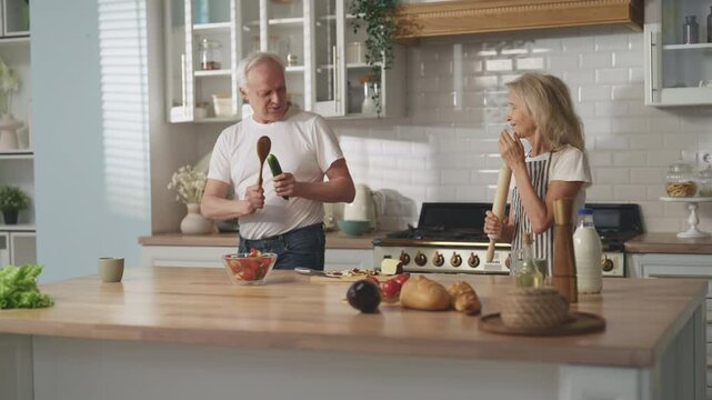 Funny aged spouses singing songs in home kitchen during cooking dinner, portrait . Happy mature man and woman having fun and fool around, weekend at home, lady holding rolling pin like a microphone