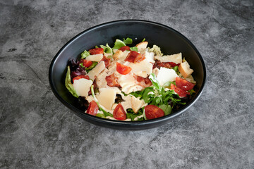 A black bowl of salad with tomatoes and cheese. The bowl is on a grey surface