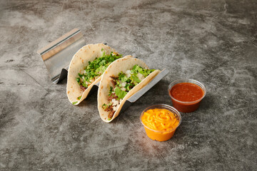 Two tacos with green lettuce and onions on top. A yellow container of sour cream and a red container of salsa