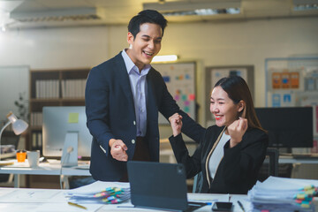 Two happy Asian businesspeople celebrating their success together, smiling and looking at a laptop in a modern office environment