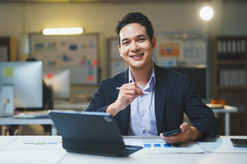 Portrait of a smiling Asian businessman working diligently with a tablet and smartphone in a modern office setting during the night