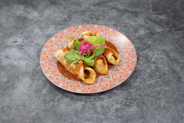 A plate of food with three tortillas on it. The plate is on a grey surface. The tortillas are covered in sauce and topped with lettuce