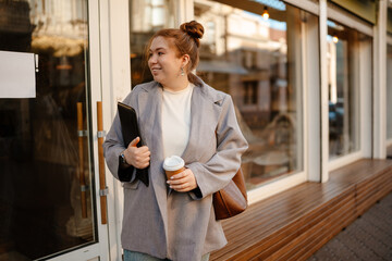 Female owner standing and smiling while looking away and holding a cup and computer tablet, on the street near the cafe