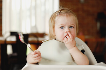 Infant girl or toddler sitting on high chair. Eating food by herself using spoon