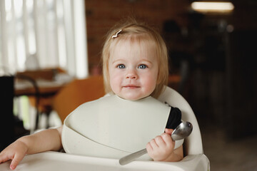 Infant girl or toddler sitting on high chair. Eating food by herself using spoon