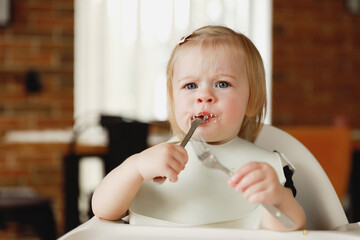 Infant girl or toddler sitting on high chair. Eating food by herself using spoon