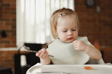 Infant girl or toddler sitting on high chair. Eating food by herself using spoon
