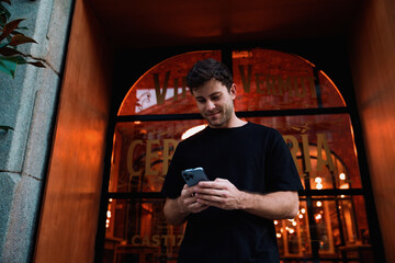 Happy young man smiles while using smartphone in front of vintage-style bar, enjoying peaceful city moment during golden hour with classic urban charm