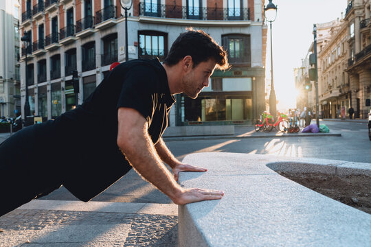 Smiling athletic man doing push-ups on city bench at sunrise, enjoying outdoor fitness routine in calm urban atmosphere, staying fit and positive