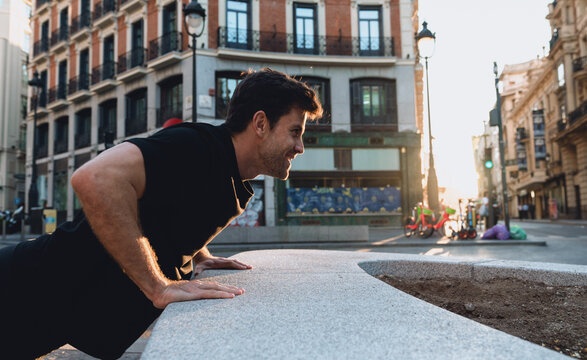 Smiling athletic man doing push-ups on city bench at sunrise, enjoying outdoor fitness routine in calm urban atmosphere, staying fit and positive