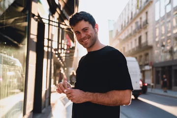 Fotobehang Backlit portrait of a modern young man with earbuds, standing confidently in sunlight flare, immersed in audiobook or podcast, surrounded by city traffic and reflections © BullRun