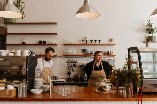 A woman owner and a man employee are standing behind the bar and smiling while putting on their aprons in a cafe