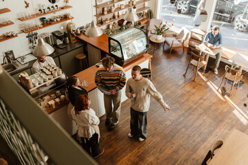 Two male customers stand and hold laptops while talking to a female employee and a female owner standing across from them, in a cafe
