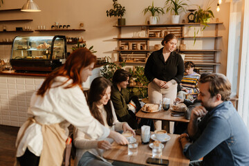 A female owner stands and talks to customers while a female employee brings coffee to a table next to her, in a cafe