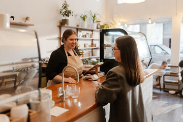 A female owner stands behind the bar and smiles as she takes an order from a female customer standing across from her in a cafe