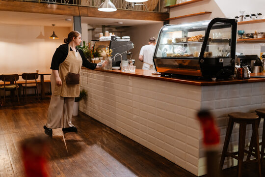 A female owner stands at the bar and holds a vase while a male employee stands next to her, in a cafe