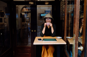 Smiling young Asian woman enjoys coffee break in cozy vintage cafe, wearing bucket hat and glasses, seated at table with smartphone and tablet, reflecting relaxed travel lifestyle