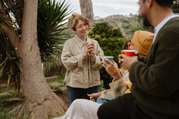 A woman stands and smiles while looking at a group of three friends sitting across from her, holding cups, outdoors