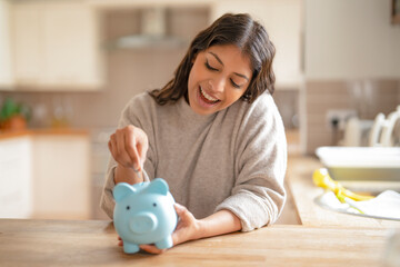 Young woman joyfully saving money in a blue piggy bank at a bright kitchen table with natural light...