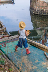 Little fisherman boy wearing straw hat on wooden boat near harbor