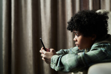 Child engrossed with smartphone, reclining on couch in cozy home environment, showing focus and...