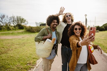 A group of three friends stand and laugh while posing and looking at a phone held by one of them,...