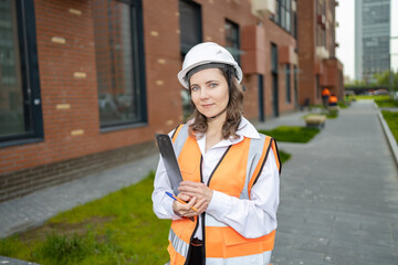 Fototapeta premium A business woman engineer in a white helmet, white shirt and orange vest against the background of new buildings, holding a tablet with documents in her hands