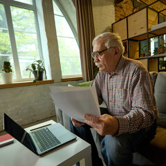 Older man with glasses concentrating on printed papers and laptop during home learning session in...
