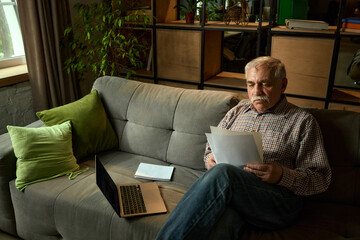 Fototapeta premium Senior man lying back on sofa and reading papers next to laptop and notebook in cozy living room. Comfortable education for seniors. Concept of elderly lifestyle, education, knowledge.
